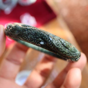 May include: Dark green jade bracelet with detailed dragon carving. The bracelet is a deep forest green colour and features detailed scales and a dragon head. The jade is polished and has a smooth, reflective surface. The bracelet is a circular shape.