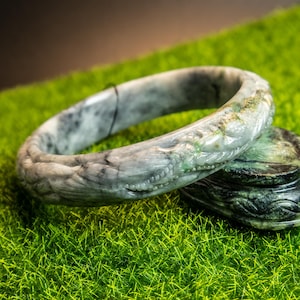 May include: A carved jade bangle with a white and green marbled pattern. The bangle is intricately carved with a dragon design.