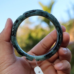 May include: A dark green jade bangle with carved detailing. The bracelet is held up against a blurred background of green and blue. The jade has a translucent quality, and the carving appears to be of a floral or organic design.