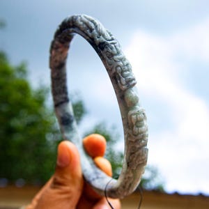May include: A carved jade bangle bracelet with a detailed relief design. The bracelet is a mix of grey and green hues, held against a backdrop of a cloudy sky. The intricate carvings depict figures and patterns.
