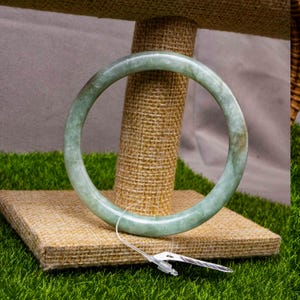 Könnte beinhalten: Ein hellgrünes Jade-Armreif-Armband. Das runde Armband hat eine glatte, polierte Oberfläche. Der Jade hat eine durchscheinende Qualität mit Farbvariationen. Das Armband wird auf einem mit Jute bedeckten Ständer vor grünem Hintergrund präsentiert.