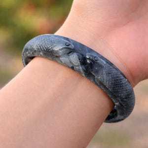May include: A dark grey jade bangle bracelet featuring two carved snake figures. The snakes have detailed scales and facial features. The bracelet is worn on a wrist, highlighting the jewellery's design and craftsmanship.
