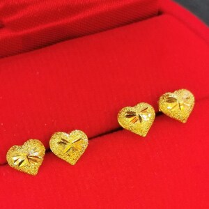May include: Three gold heart-shaped earrings on a red velvet background. The earrings are small and have a textured surface.
