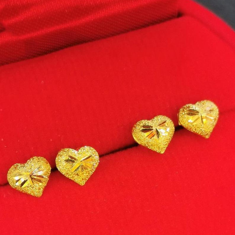 May include: Three gold heart-shaped earrings on a red velvet background. The earrings are small and have a textured surface.