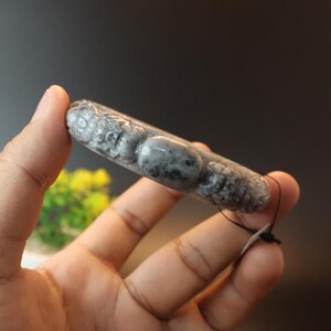 May include: A grey and black carved jade bracelet with a black string attached. The bracelet has a detailed, textured surface with a pattern of rounded shapes. The jade is held in a hand against a dark background.
