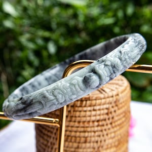 May include: A grey and green jade bangle bracelet with detailed carvings. The bracelet is presented on a gold-coloured stand, highlighting its design and colour variations. The background is blurred, focusing attention on the bracelet's craftsmanship.