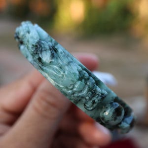 May include: A carved jade bracelet in shades of green and black. The bracelet features a detailed relief carving of a dragon. The jade is polished and has a smooth, reflective surface. The bracelet is held in a hand.