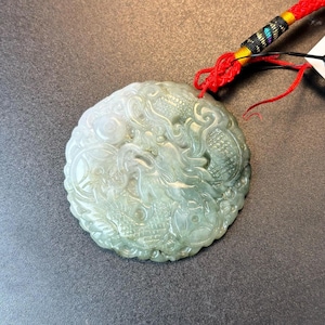 May include: A jade pendant featuring an intricate carving of a dragon. The pendant is attached to a red and black cord.