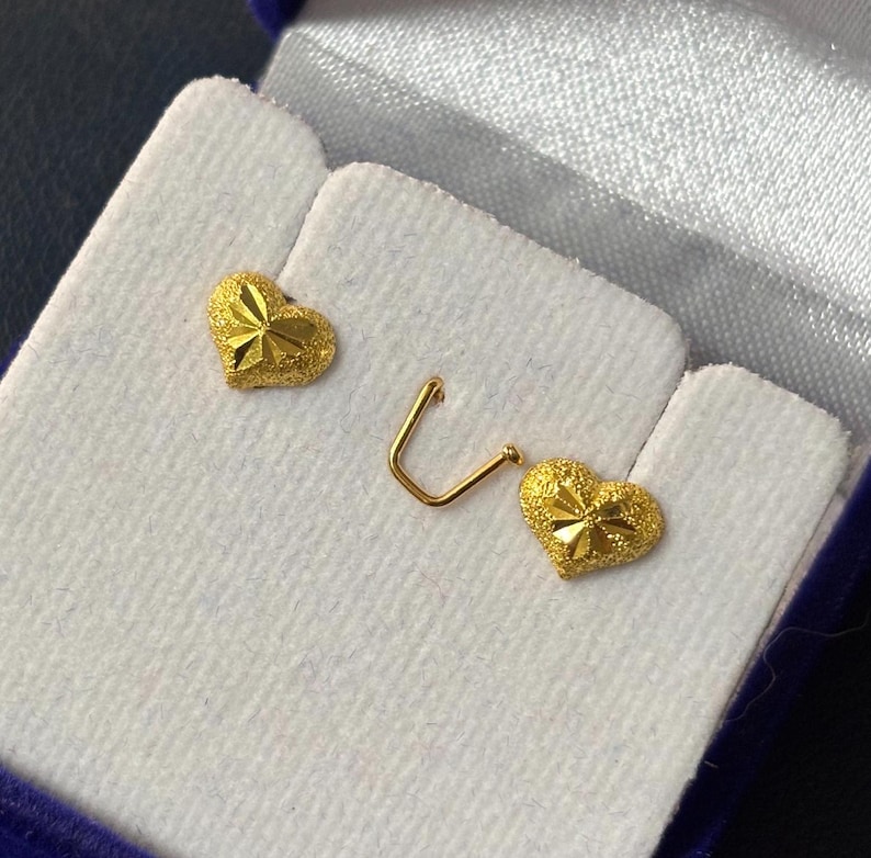 May include: Two gold heart-shaped earrings with a textured surface. The earrings are displayed in a blue velvet box.