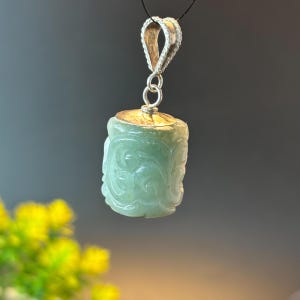 May include: A light green jade pendant with detailed floral carvings. The pendant features a silver-coloured bail and a gold-coloured cap. It hangs from a black cord, highlighting the craftsmanship and natural beauty of the stone.
