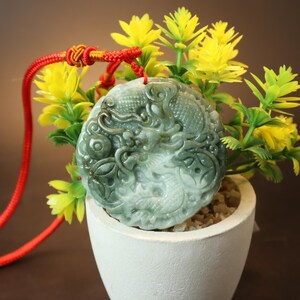 May include: A jade pendant featuring a carved dragon design, suspended from a red cord. The pendant is green and white with intricate details. The pendant is shown against a backdrop of yellow flowers.