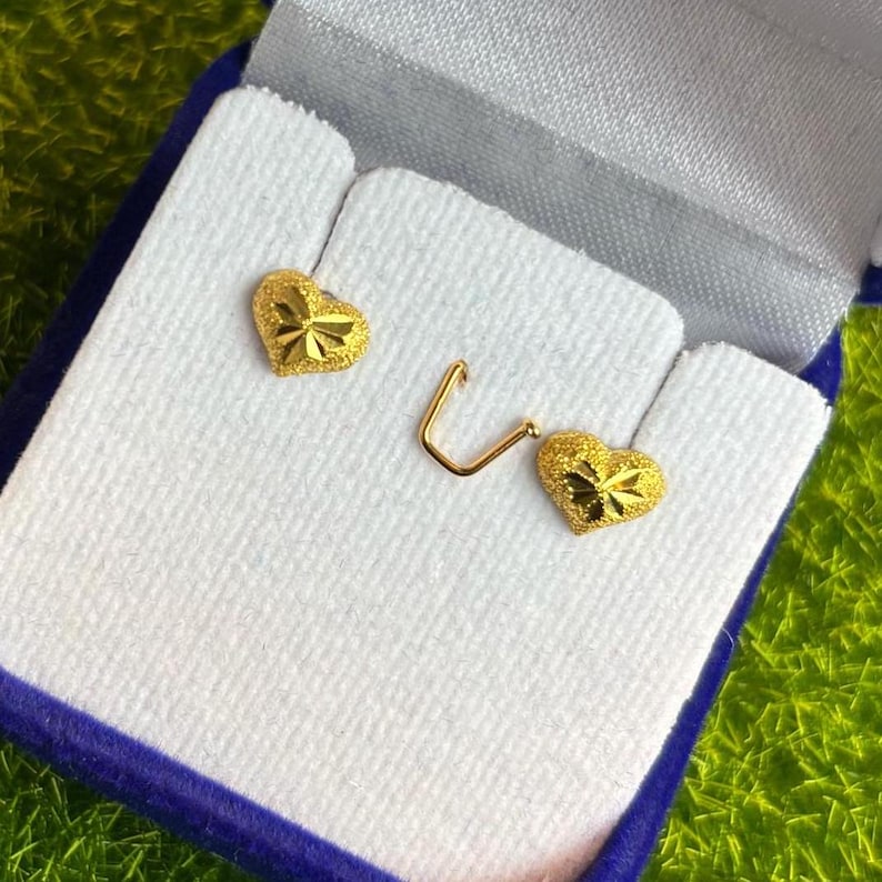 May include: A pair of gold heart-shaped earrings in a white box with a blue velvet lining. The earrings have a textured surface.