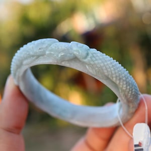 May include: A pale green jade bracelet with a dragon motif. The bracelet displays a carved dragon head and a textured, scale-like pattern. The jade has a translucent quality, with a subtle green hue.