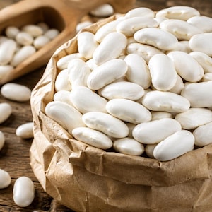 Puede incluir: Una bolsa de papel marrón llena de numerosos frijoles blancos lisos y ovalados. Una pala de madera con más frijoles blancos descansa cerca. Los frijoles tienen un acabado mate y la bolsa presenta una textura arrugada en la base.