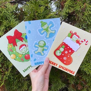 May include: Three Christmas cards with festive illustrations. One card features a wreath with a bow, another shows snow angels, and the last depicts a stocking with a candy cane. The cards have the words "Merry", "Happy", and "Merry Smiskmas".