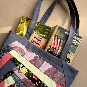 May include: A gray tote bag with patchwork fabric in shades of pink, green, and gray. The bag has two long handles and is filled with three books. The books have titles that include "Jack London", "Tre Inimi", and "fran".