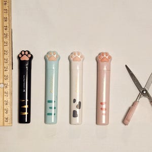 May include: A collection of cat paw-shaped scissors and pen-like containers. The containers are black, light blue, white, and pink. The scissors have pink handles. A wooden ruler is on the left side. The text "Cat Paw Scissors" is on the black container.
