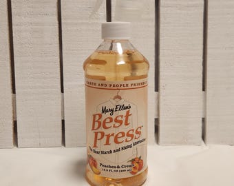 May include: A clear plastic spray bottle filled with a light amber liquid. The label reads "Mary Ellen's Best Press" with "Peaches & Cream" flavor. The bottle contains 16.9 fl oz (499 ml) of clear starch and sizing alternative.