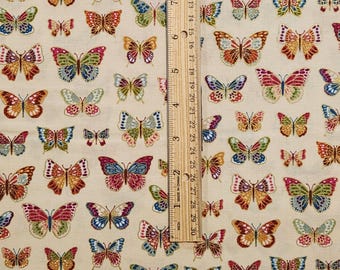 May include: Cream-colored fabric patterned with numerous colorful butterflies. The butterflies vary in size and feature a range of colors including red, blue, green, and gold. A ruler is placed vertically on the fabric, showing inch measurements.