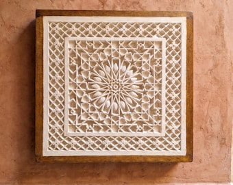 Könnte beinhalten: Ein quadratisches Wandpaneel mit einem komplexen geometrischen Muster aus weißem, geschnitztem Gips und einem zentralen Sternmotiv, eingefasst in einen dunklen Holzrahmen. Das Design besteht aus sich wiederholenden Gitter- und Blumenelementen.