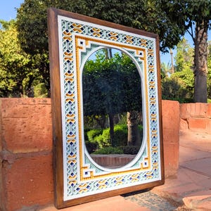 May include: Rectangular decorative mirror with an oval center. The frame is wood-toned with a detailed border of blue, yellow, and white geometric patterns. The mirror reflects a garden scene, adding depth and visual interest to the design.