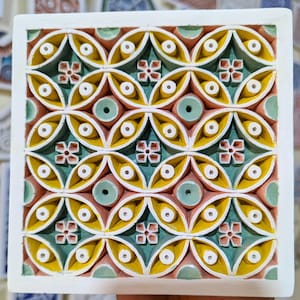 Peut inclure: Un carreau de céramique carré avec un motif géométrique répétitif dans les tons de jaune, vert, rose et blanc. Le carreau a un design en relief avec de petits cercles et des formes de fleurs.