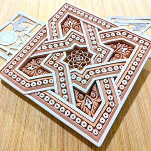 May include: A white and brown ceramic tile with an intricate geometric design. The tile features a star shape with a smaller star in the center. The tile is surrounded by a border of small circles.