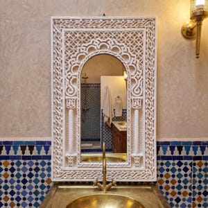 May include: A rectangular wall mirror featuring an intricate, carved off-white frame with geometric and interlacing patterns. The mirror is positioned above a metallic gold sink basin and a matching gold faucet, reflecting a tiled bathroom interior.