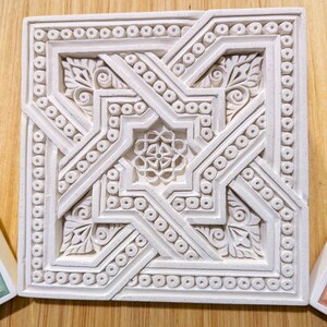 May include: A white ceramic tile with an intricate geometric design. The design features a central star shape surrounded by smaller stars and floral motifs. The tile is bordered by a row of small circles.