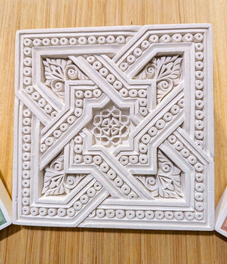 May include: A white ceramic tile with an intricate geometric design. The design features a central star shape surrounded by smaller stars and floral motifs. The tile is bordered by a row of small circles.