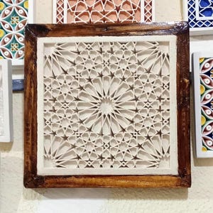 May include: A square wall art piece featuring a cream-colored, multi-layered geometric lattice pattern with intricate star and polygon shapes. The central design is encased within a dark, polished wooden frame with a visible grain texture.