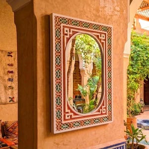 May include: A rectangular wall mirror featuring an oval reflective centre surrounded by a wide, flat frame. The frame is decorated with a repeating geometric pattern in shades of terracotta, cream, and dark green, set against a light wall.