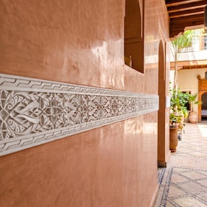 May include: A peach-colored wall featuring a horizontal band of intricate white plaster relief carving with geometric and floral patterns. The wall includes an arched opening and leads to an outdoor courtyard with tiled flooring and greenery.