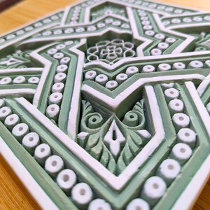 May include: A green and white ceramic tile with an intricate geometric pattern. The tile features a star shape in the center, surrounded by a series of concentric circles and lines.