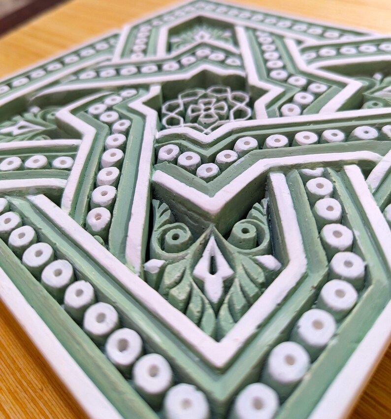 May include: A green and white ceramic tile with an intricate geometric pattern. The tile features a star shape in the center, surrounded by a series of concentric circles and lines.