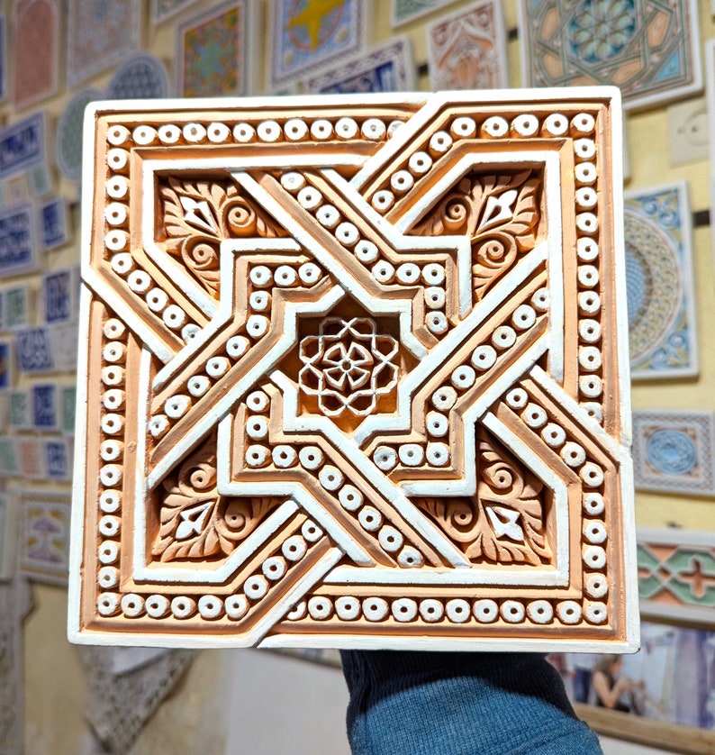 May include: A square ceramic tile with a white and orange geometric design. The tile has a raised, intricate pattern with a central star shape.