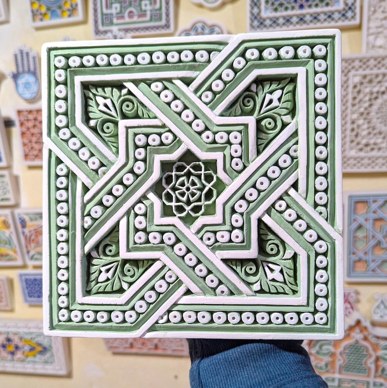 May include: A square, green and white ceramic tile with an intricate geometric design. The tile features a central star shape surrounded by a series of interlocking lines and circles.
