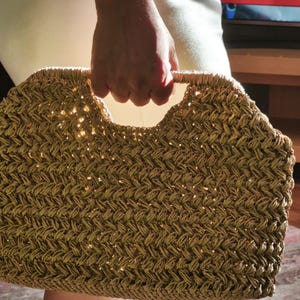 May include: A woven straw handbag with a rectangular shape and a curved handle. The bag has a textured pattern and a natural, light brown color. The handle is a lighter shade of brown.