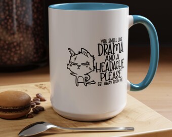 Puede incluir: Taza de cerámica blanca con borde y asa azules. La taza presenta una ilustración en blanco y negro de un gato con el texto "You smell like drama and a headache please get away from me".