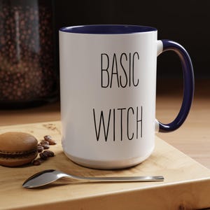 May include: White ceramic mug with a blue rim and handle. The mug has the text "BASIC WITCH" printed in black.