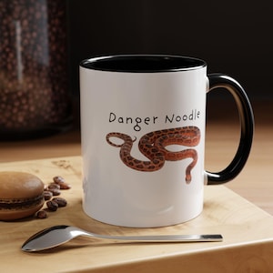 May include: White ceramic mug with a black interior and handle. The mug features the text "Danger Noodle" above a brown and tan snake illustration. A spoon and a pastry are in the foreground.