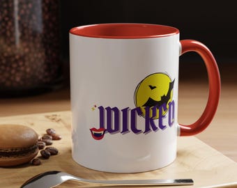 Puede incluir: Una taza de cerámica blanca con un borde y un asa rojos. La taza tiene un gráfico de una luna amarilla con un murciélago volando frente a ella. El texto "WICKED" está en letras moradas con un gráfico de boca roja debajo.