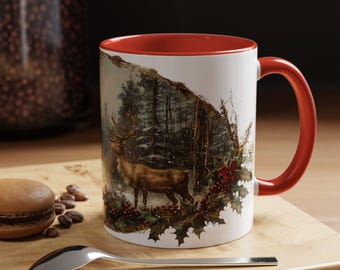 May include: White ceramic mug with a red interior and handle. The mug features a detailed illustration of a deer in a snowy forest scene with holly and berries. A spoon and a macaron are on a wooden surface.