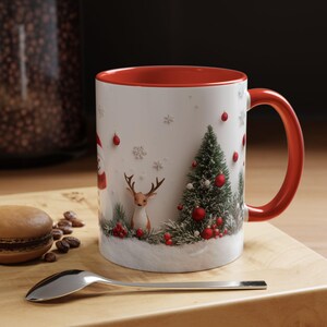 Puede incluir: Taza de cerámica blanca con borde y asa rojos. La taza presenta una escena de invierno con un muñeco de nieve, un ciervo, un árbol de Navidad y adornos rojos. El fondo es blanco con copos de nieve.