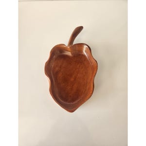 Puede incluir: Un cuenco de madera con forma de hoja y un tallo. El cuenco está hecho de madera marrón oscuro y tiene un acabado liso y pulido. El cuenco está vacío y descansa sobre una superficie blanca. El cuenco mide unos 20 cm de largo.