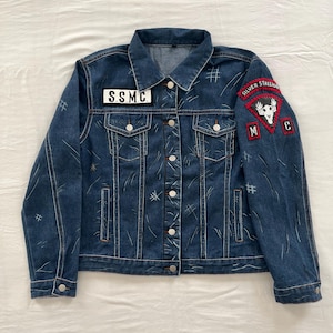 May include: A blue denim jacket with white stitching and several patches. The patches include "S.S.M.C." and "SILVER STALLION" with a skull and crossbones graphic.
