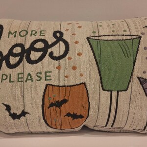 May include: Halloween-themed decorative pillow with the words "MORE BOOS PLEASE" in black and green. The pillow features a green cocktail glass, an orange pumpkin, bats, and a spider. The background has a wood-like texture.
