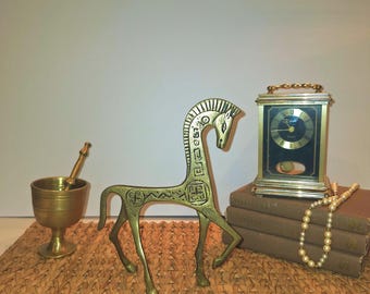 May include: A brass horse figurine with an elongated neck and decorative etchings stands on a woven mat. A brass mortar and pestle, a vintage clock, and a strand of pearls are also visible.