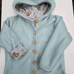 Könnte beinhalten: Hellblaue Kapuzenjacke mit Holzknöpfen. Die Innenseite der Kapuze und der Manschetten ist mit einem grauen Stoff mit einem sich wiederholenden Muster aus braunen Zeichentricktieren und Blättern versehen. Die Jacke hat eine Knopfleiste und ein gemütliches Design.