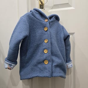 May include: A blue hooded jacket with four wooden buttons. The jacket has long sleeves and a patterned lining visible at the cuffs. The jacket is hanging on a white hanger.
